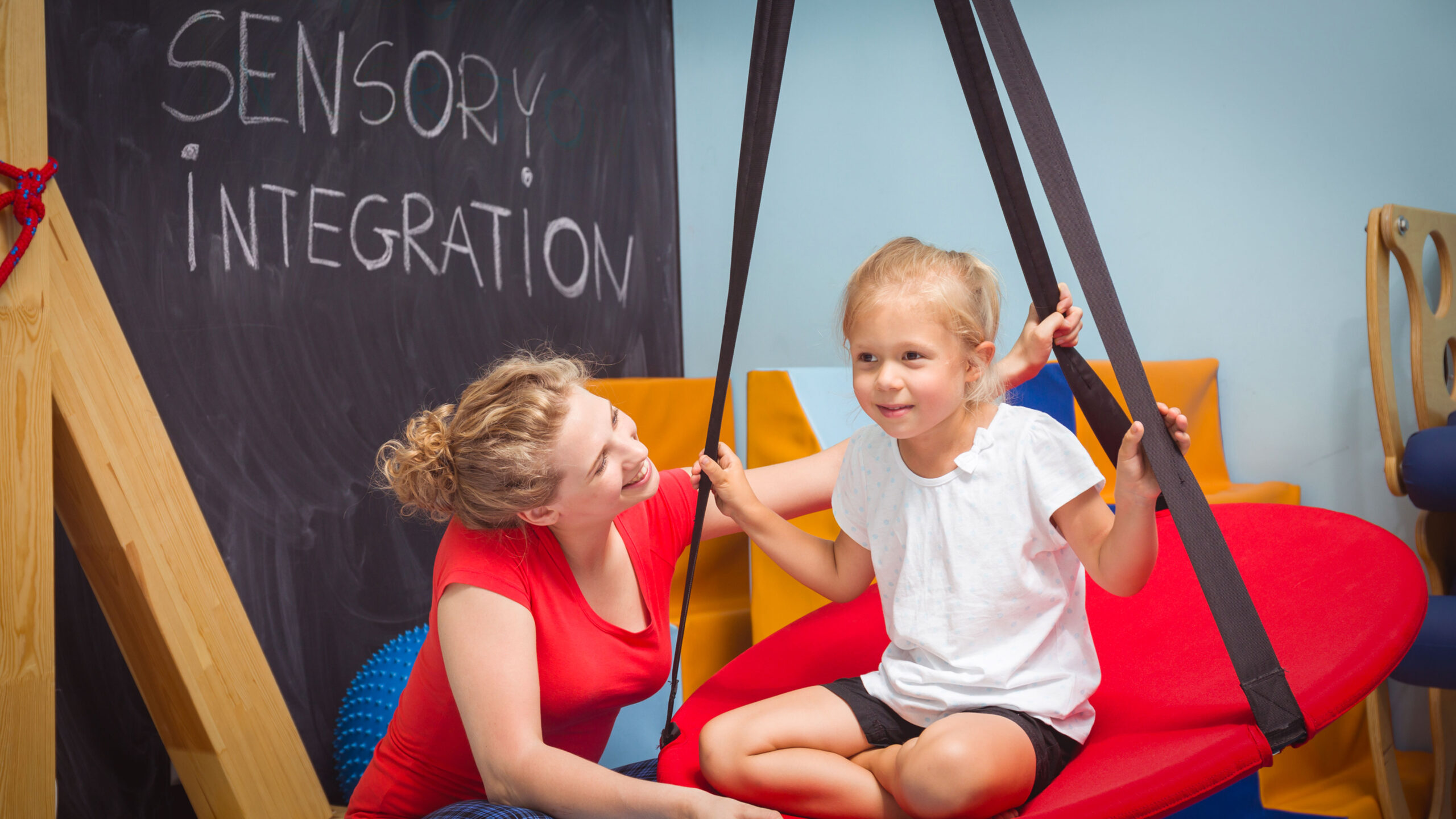 A girl swinging on a sensory swing guided by a therapist.